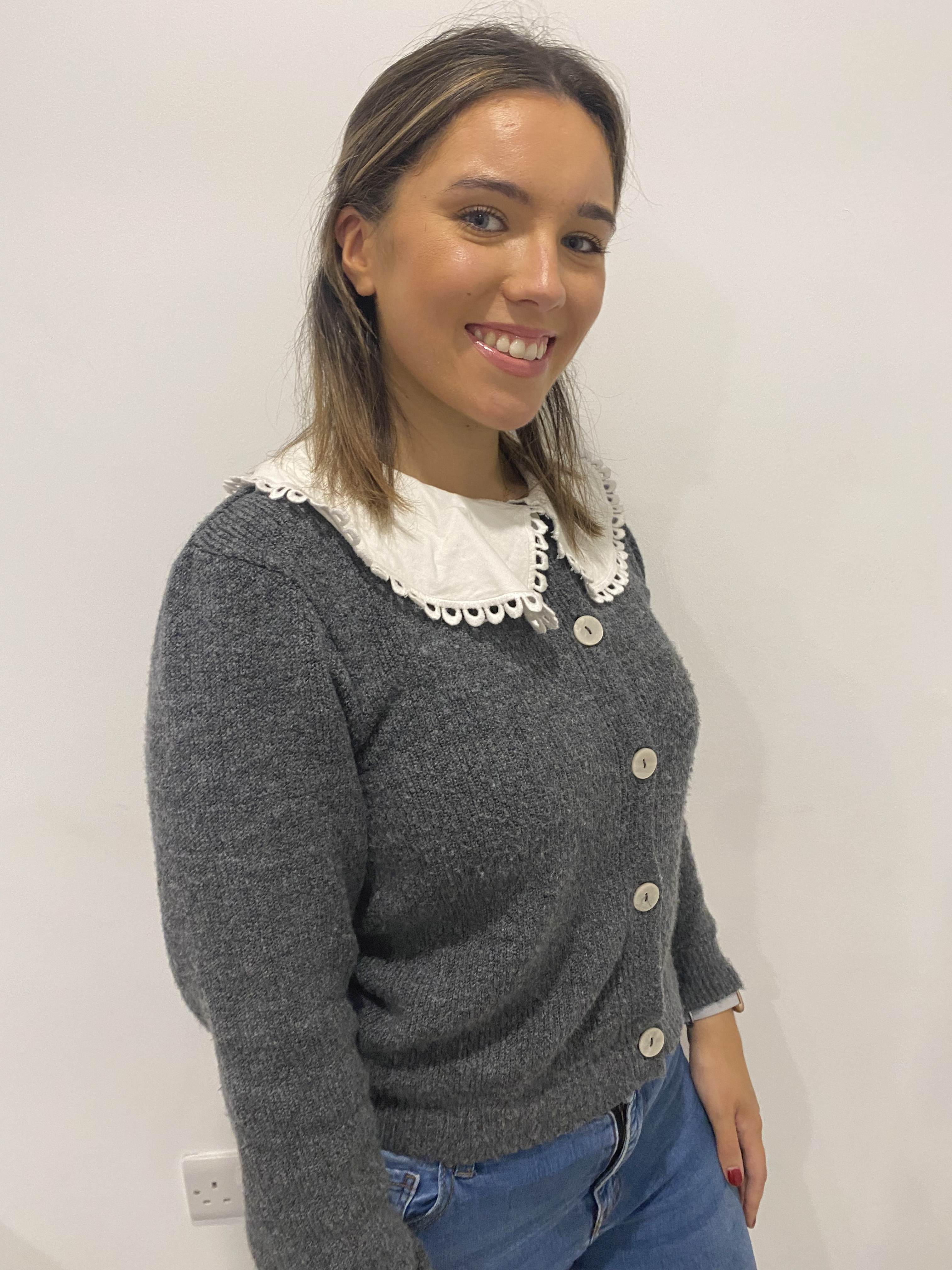 The image shows Amy Sweeney, a family support worker with Enable Ireland, standing and smiling. Amy is dressed in a grey cardigan with blue jeans. 